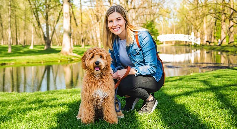 Smiling woman in a denim jacket crouching beside a fluffy brown dog on a sunny lawn by a pond.