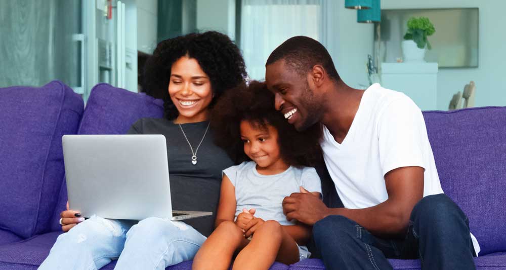 Family of three sitting on a purple couch looking at a laptop.