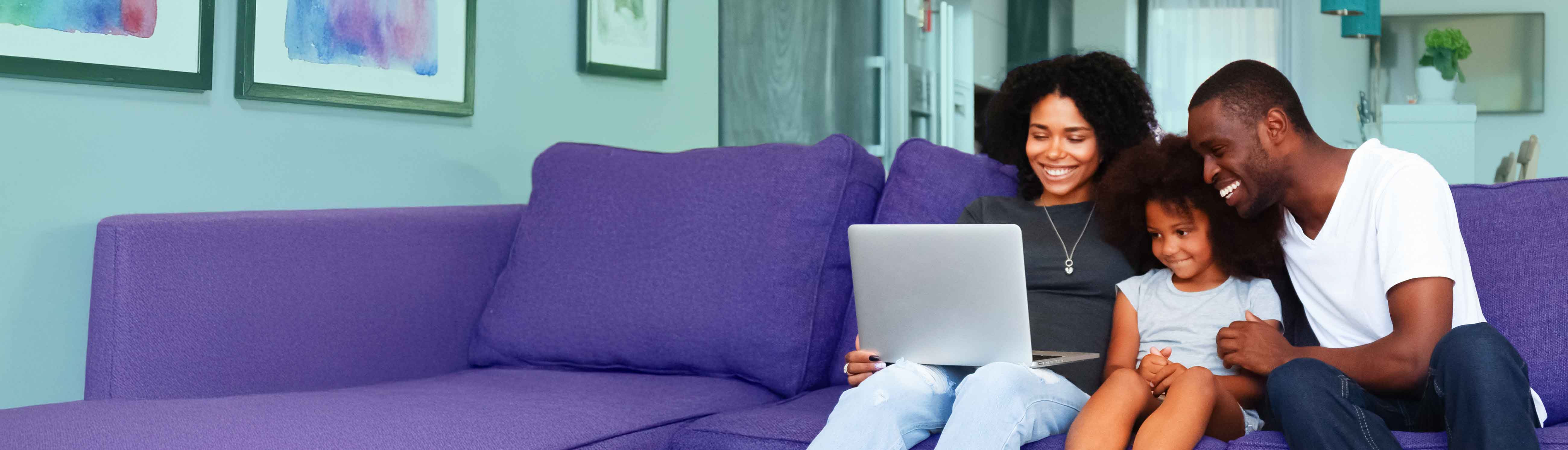 Family of three sitting on a purple couch looking at a laptop.